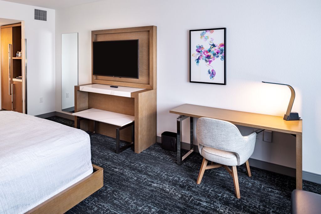 A hotel room with a bed, a desk, a chair, a television, and a framed artwork on the wall.