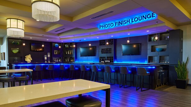 A bar area with a sign reading 'PHOTO FINISH LOUNGE,' bar stools, a bar counter, and a television mounted on the wall. - Prairie Meadows Hotel (Altoona)