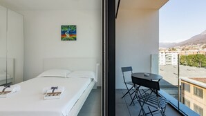 A hotel room with a bed, a painting, a balcony with a table and chairs, and a view of buildings and mountains. - {Suite 608} Gym, Business Room, Modern, Relax (Lugano)