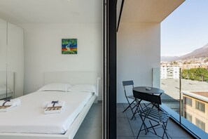 Quarto de hotel com uma cama, um quadro, varanda com mesa e cadeiras, e vista para edifícios e montanhas.