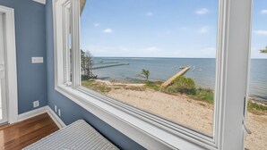 Een kamer met een groot raam met uitzicht op een strand, een houten vloer en een witte deur.