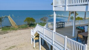 Een pand aan het strand met een houten terras, buitenmeubels en een trap naar een balkon.