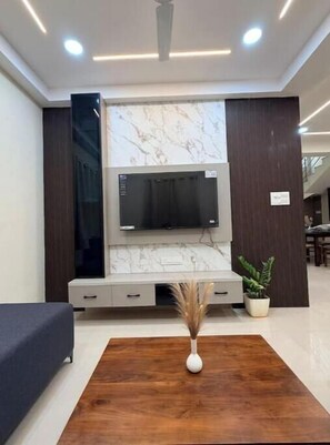 Un salón moderno con un televisor de pantalla plana grande montado en una pared de mármol, un sofá oscuro, una mesa de centro de madera con un florero y una planta en maceta.