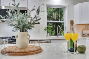 Une cuisine moderne avec des meubles blancs, une hotte de cuisine en acier inoxydable, une fenêtre donnant sur un espace verdoyant, une plante en pot et une bouteille de jus d’orange.