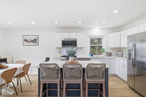 Une cuisine moderne avec des meubles blancs, un réfrigérateur en acier inoxydable et un coin repas comprenant des chaises en osier et une table blanche.