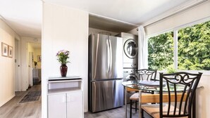 A modern kitchen with stainless steel appliances, a dining area with a glass table and chairs, and a view of greenery outside.