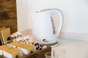 Ein weißes Wasserkocher mit Griff und Bedienfeld, eine Holzkiste mit Kaffee-Kapseln und ein Glas auf einer weißen Arbeitsplatte.