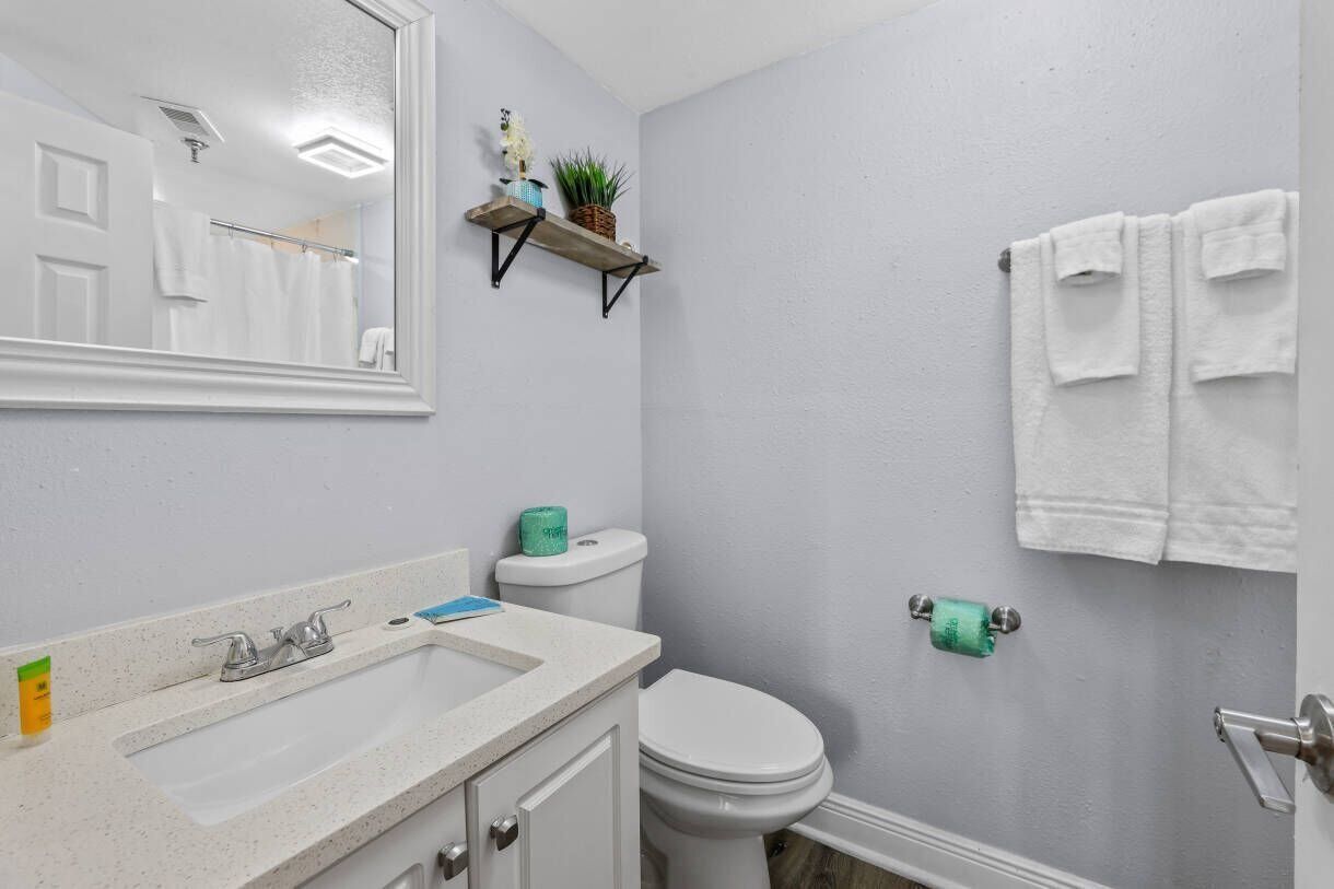 Een badkamer met een wit wastafelmeubel, toilet en spiegel. Handdoeken hangen aan de muur en er is een plankje met een plant en een klein voorwerp.