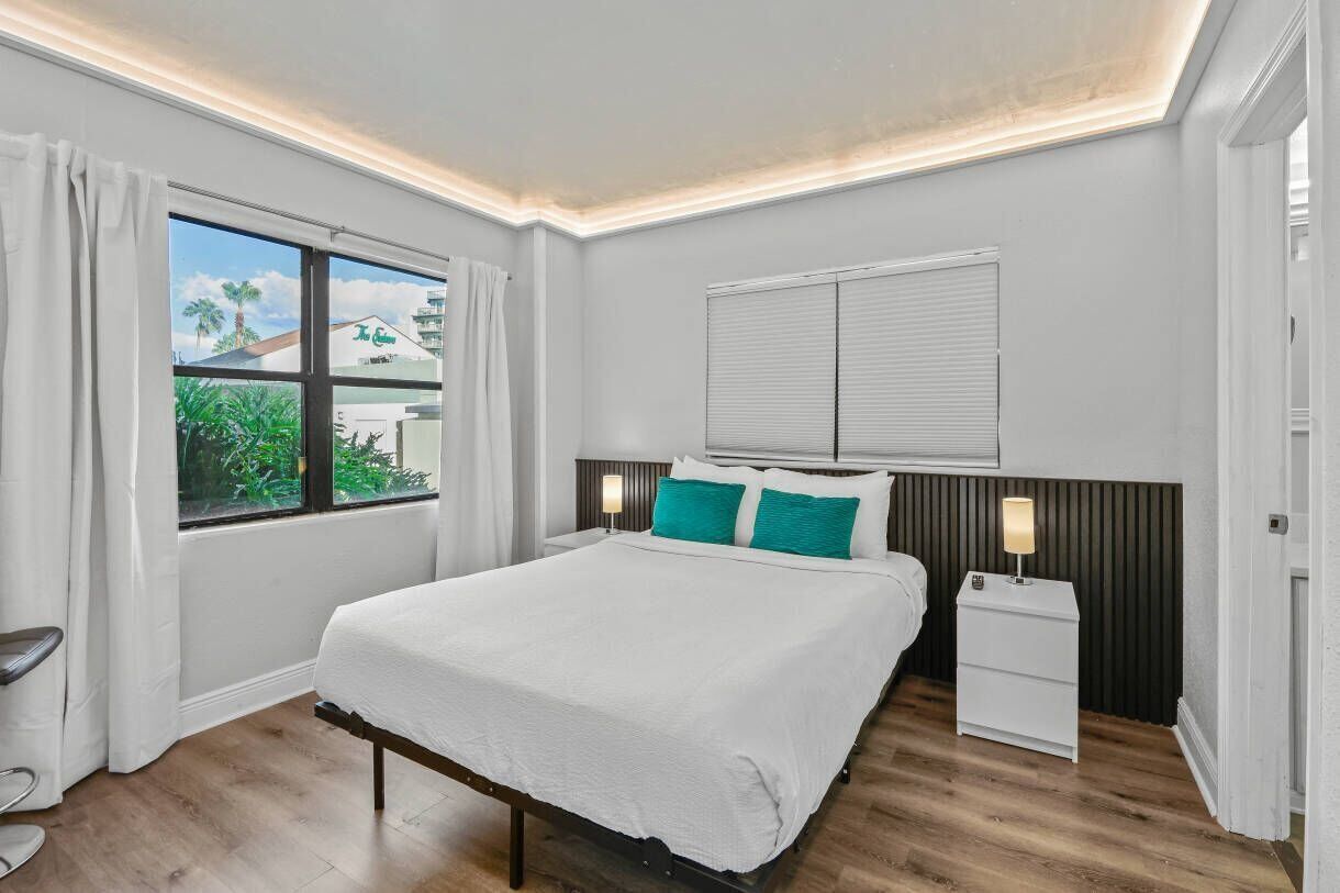 A hotel room with a bed, a nightstand, a window with curtains, and a view of greenery outside.
