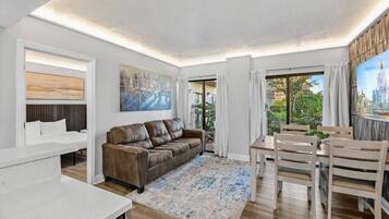 A modern living room with a brown leather sofa, a dining table with chairs, a large painting, and a view of greenery outside.