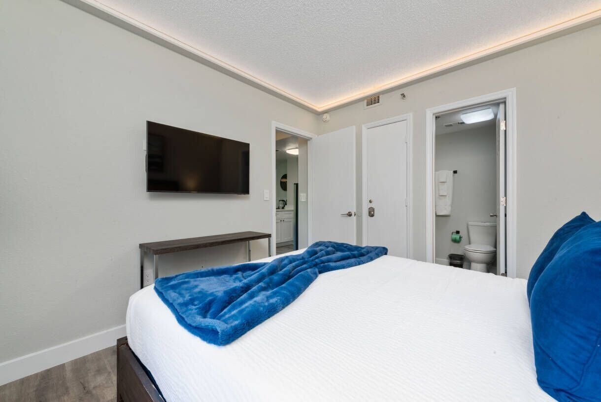 Un dormitorio con cama, televisor montado en la pared, mesita de noche y baño visible a través de una puerta abierta.