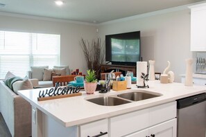 A modern kitchen with a double sink, a flat-screen TV, and a welcoming 'welcome' sign. - Beautiful Palm Coast Escape Near Beaches (Palm Coast)