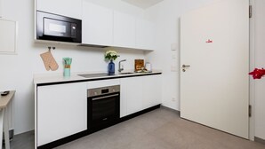 Ett kompakt kök med mikrovågsugn, ugn och diskho, en vit dörr med ett rött handtag och en liten bänkskiva med en vas med blommor.
