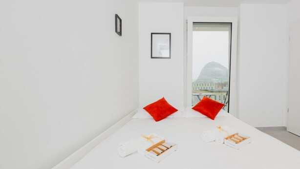 Ett minimalistiskt hotellrum med ett stort fönster, vita väggar och utsikt över byggnader.