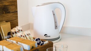 En vit, elektrisk vattenkokare med handtag och kontrollpanel, en trälåda med kaffekapslar och ett glas på en vit bänkskiva.