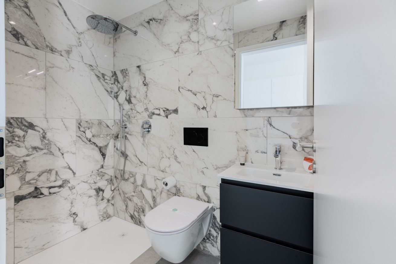 A bathroom with a marble wall, a shower, a toilet, and a sink with a mirror above.