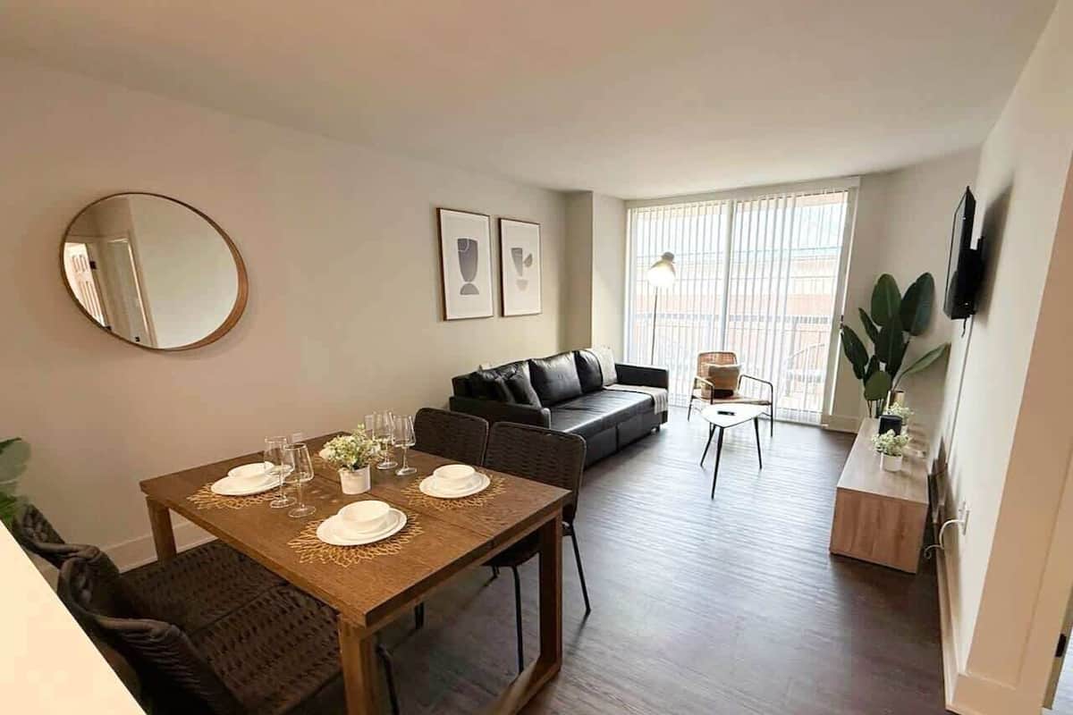Un comedor con una mesa preparada para cuatro personas, un sofá, un televisor y una ventana con persianas.