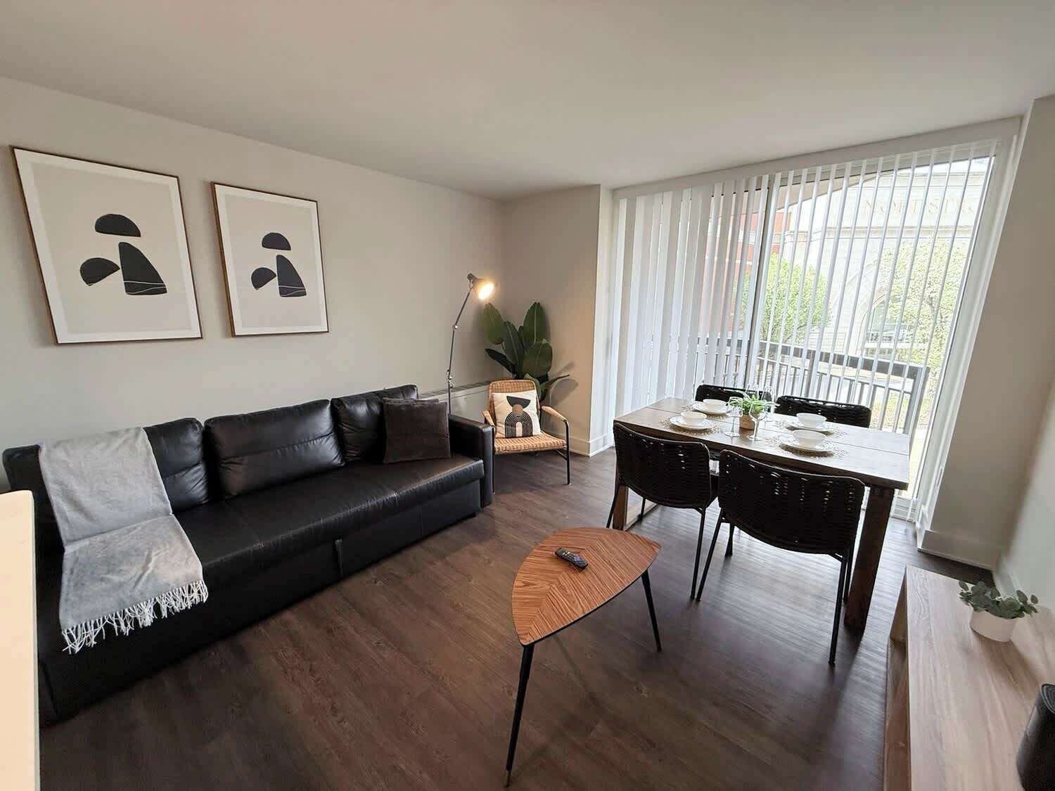 Ein modernes Wohnzimmer mit einer schwarzen Ledersofa, einem Holztisch, einem Esstisch mit Stühlen und einem großen Fenster mit Jalousien.