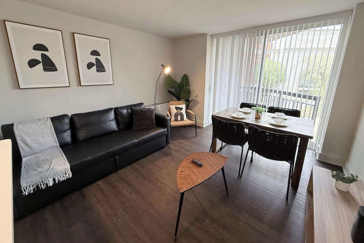 Una sala de estar moderna con un sofá de cuero negro, una mesa de centro de madera, una mesa de comedor con sillas y un amplio ventanal con persianas.