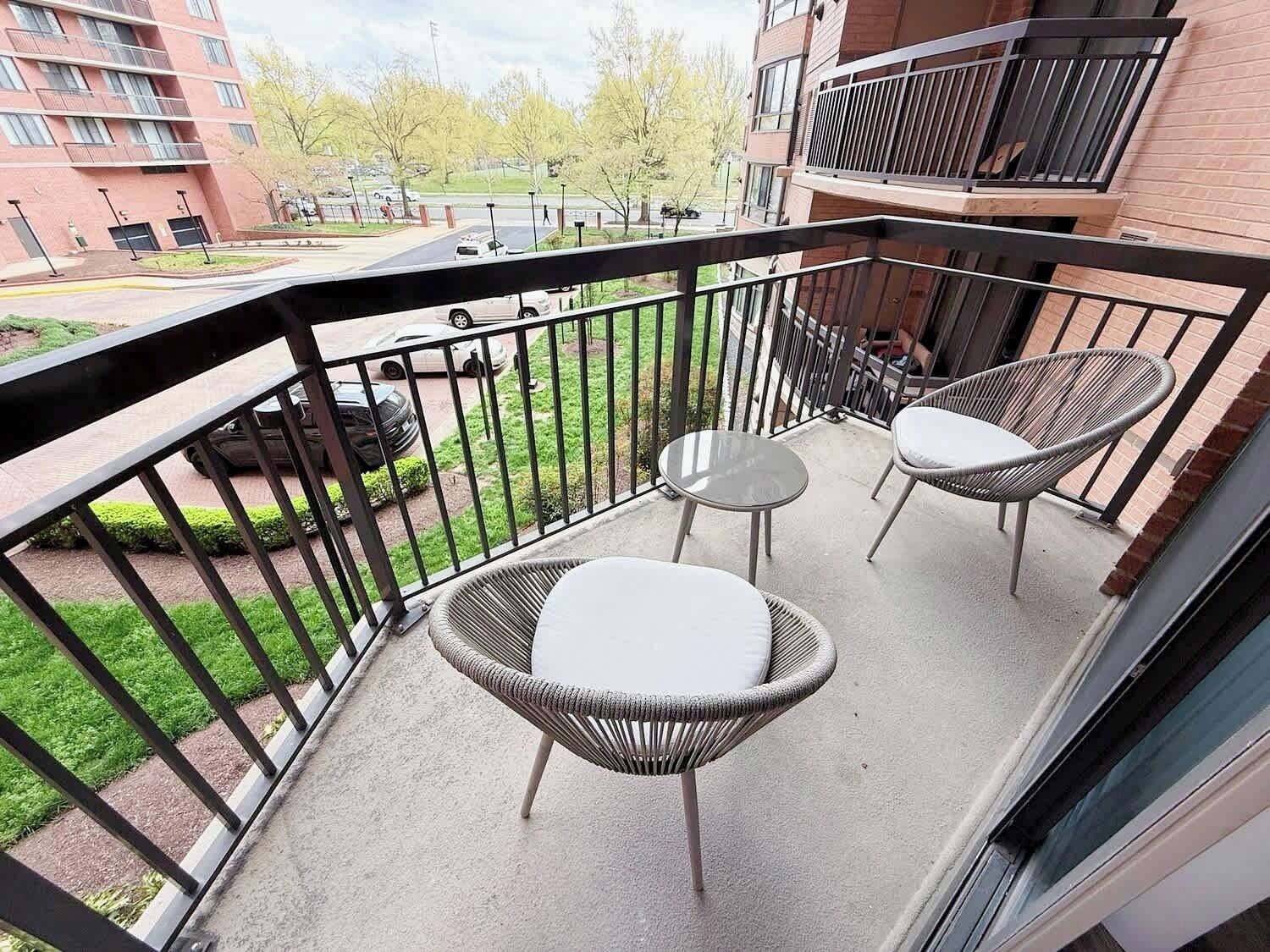 Ein Balkon mit Korbstühlen und einem kleinen Tisch, mit Blick auf einen Parkplatz und Gebäude.