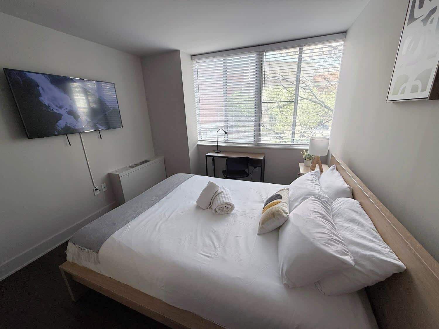 Een hotelkamer met een bed, een aan de muur bevestigde televisie, een bureau en een raam met jaloezieën.