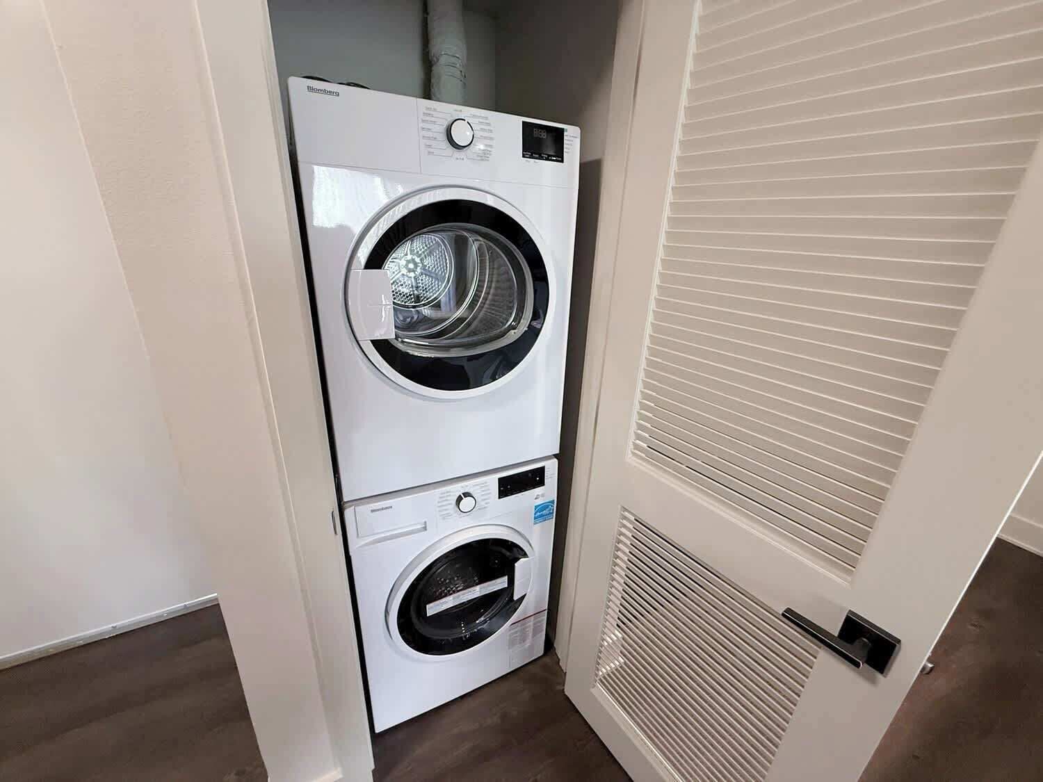A compact, white, two-door washer and dryer unit in a small laundry room with a vented door and a wooden floor.