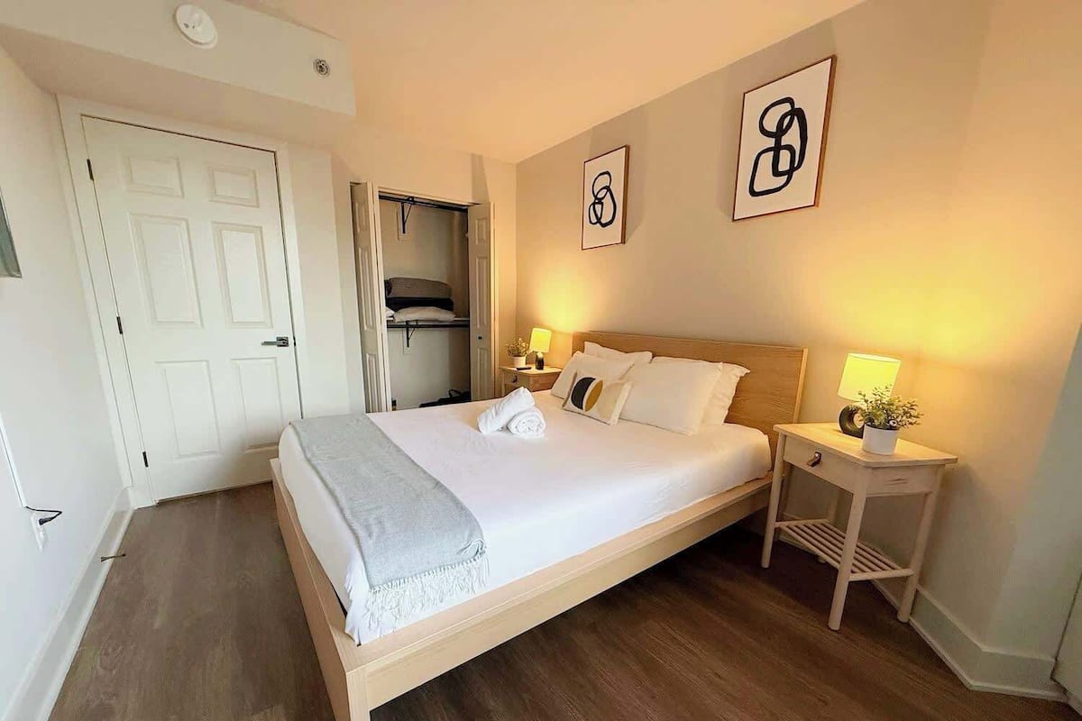 A neatly made bed with a grey blanket, a bedside table with a lamp and a plant, a closet with a hanging garment, and two abstract wall art pieces.