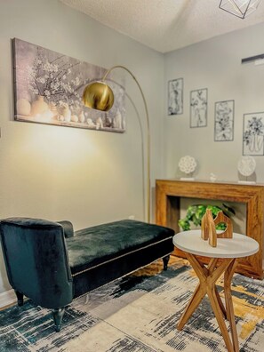 A room with a black velvet sofa, a round wooden table with two decorative items, a modern floor lamp, and a fireplace with a green plant. - 5BR Pool Home w/ Fire Pit . Sleeps 10 (Deltona)
