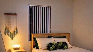 Een slaapkamer met een bed, nachtkastje, lamp en een raam met jaloezieën.