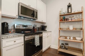 A kitchen with white cabinets, a stainless steel microwave, a stove, and a wooden shelf with kitchenware. - Gated King Condo Steps to Pier Village (St. Simons Island)