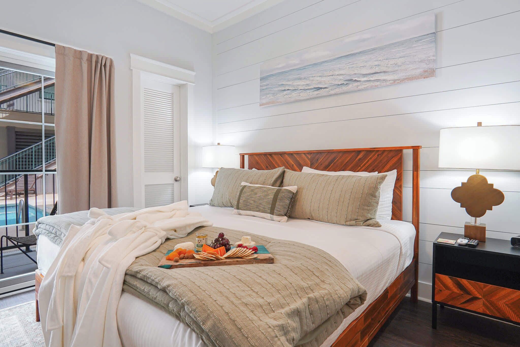 A hotel room with a bed, a bedside table with a lamp, a view of a balcony, and a painting on the wall.