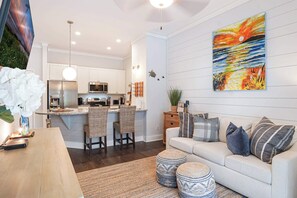A modern kitchen with white cabinets, a central island with bar stools, and a dining area with a white sofa and striped cushions. - Gated King Condo Steps to Pier Village (St. Simons Island)