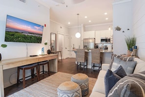 A modern living room with a large flat-screen TV, a dining area with wicker chairs, a kitchen with stainless steel appliances, and a cozy seating area with ottomans. - Gated King Condo Steps to Pier Village (St. Simons Island)