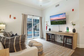 A living room with a sofa, ottoman, and a TV on the wall. - Gated King Condo Steps to Pier Village (St. Simons Island)