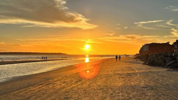 Een strand met een zonsondergang, mensen die wandelen en gebouwen op de achtergrond.