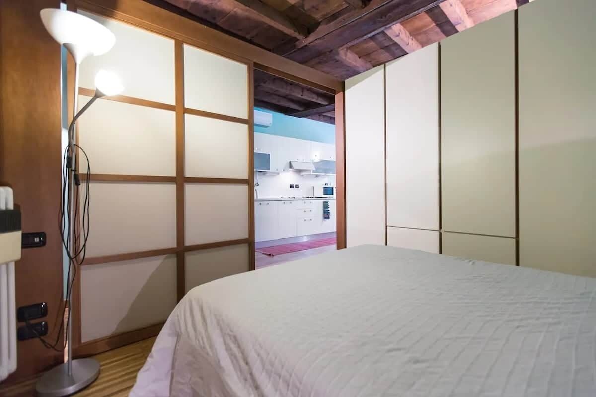 A room with a bed, a floor lamp, sliding doors, and a view of a kitchen.