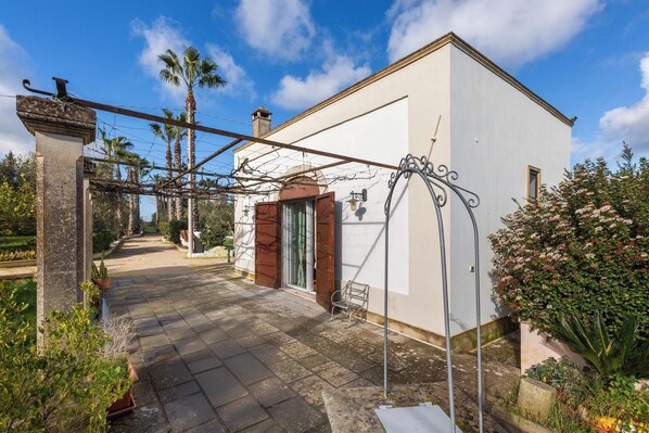 A white building with a brown door and a small window, surrounded by greenery and a paved pathway. - Le Dimore di Casina Litta - La Dependance (Cutrofiano)