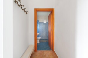 Ein enger Flur führt zu einem Badezimmer mit Toilette und Waschbecken, einem blauen Duschbereich und einem dekorativen Metallkunstwerk an der Wand.