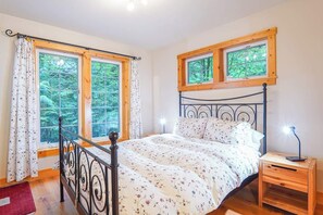 A bedroom with a bed, two windows, a nightstand, and a red rug. - Creekside Cabin • Hot Tub, Pool & Ping Pong (Deming)