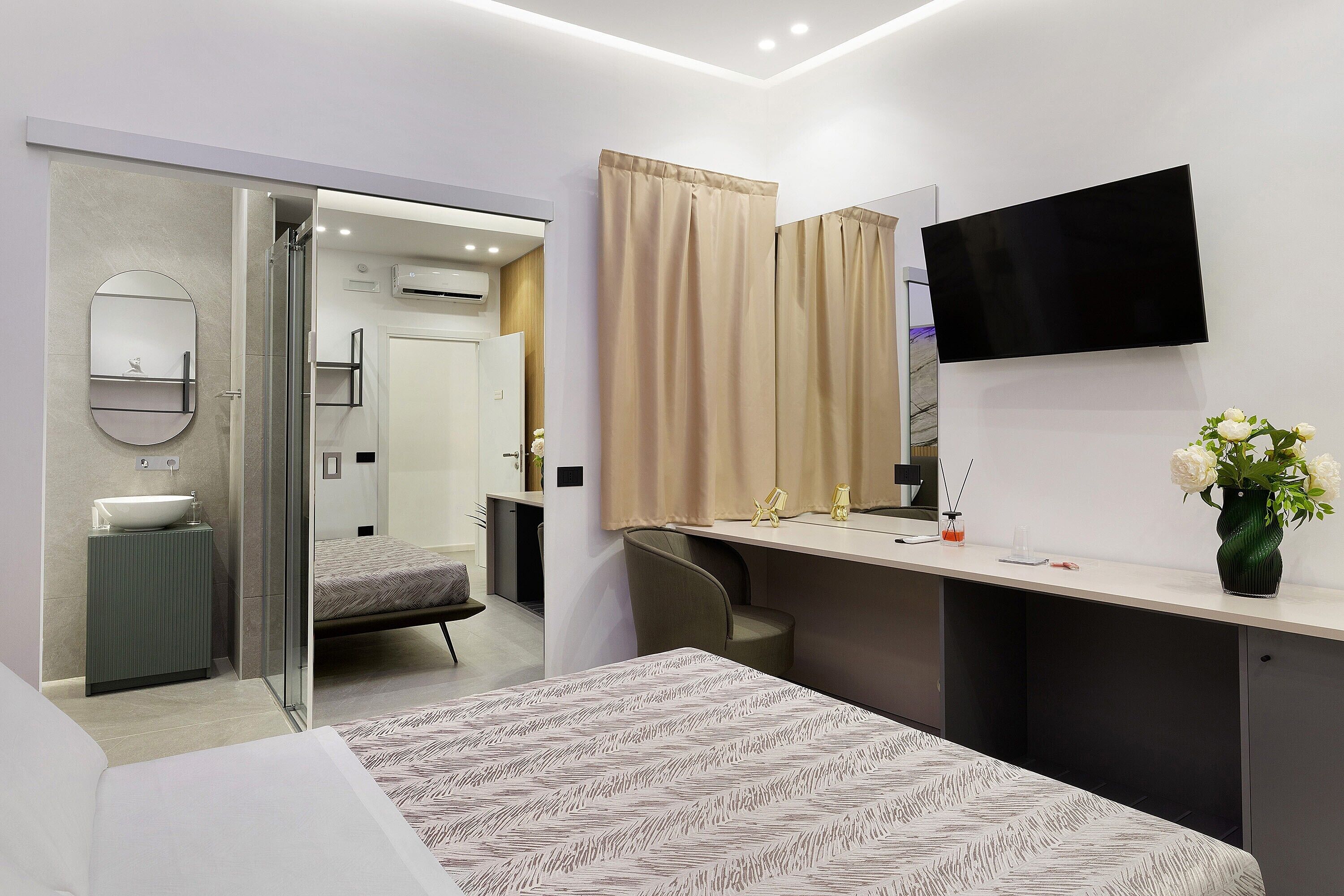A modern hotel room with a bed, a bedside table, a mirror, a TV, and a bathroom with a shower and toilet.
