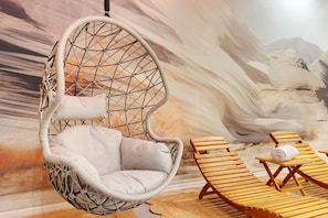 A hanging chair in a room with a desert mural, wooden lounge chairs, and a woven rug.