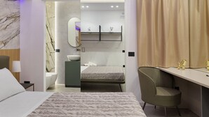 A modern hotel room with a bed, a desk, a chair, a bathroom with a marble wall, and a shower area. - Deluxe Suite with Balcony, Wi-Fi, and Air Conditioning (Partinico)