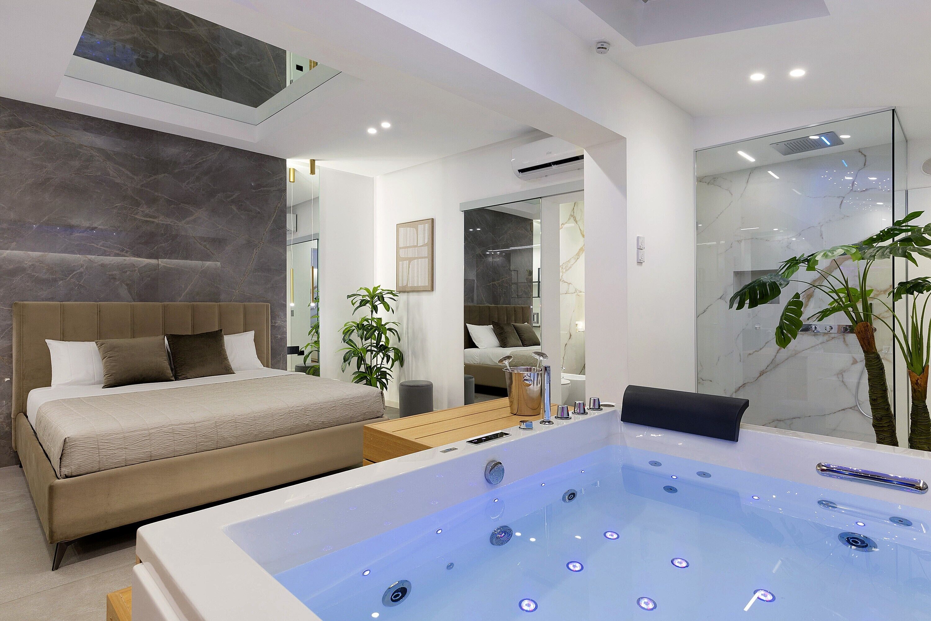 A modern bedroom with a large bed, a sitting area, a bathroom with a glass shower, and a jacuzzi.