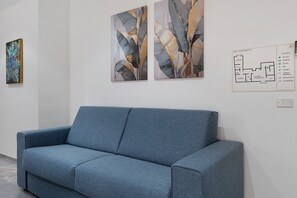 Un canapé bleu dans une pièce aux murs blancs, deux tableaux abstraits et un plan d’étage accroché au mur.