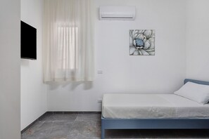 Une chambre à coucher avec un lit, une fenêtre avec des rideaux légers, un appareil de climatisation et une œuvre d’art accrochée au mur.