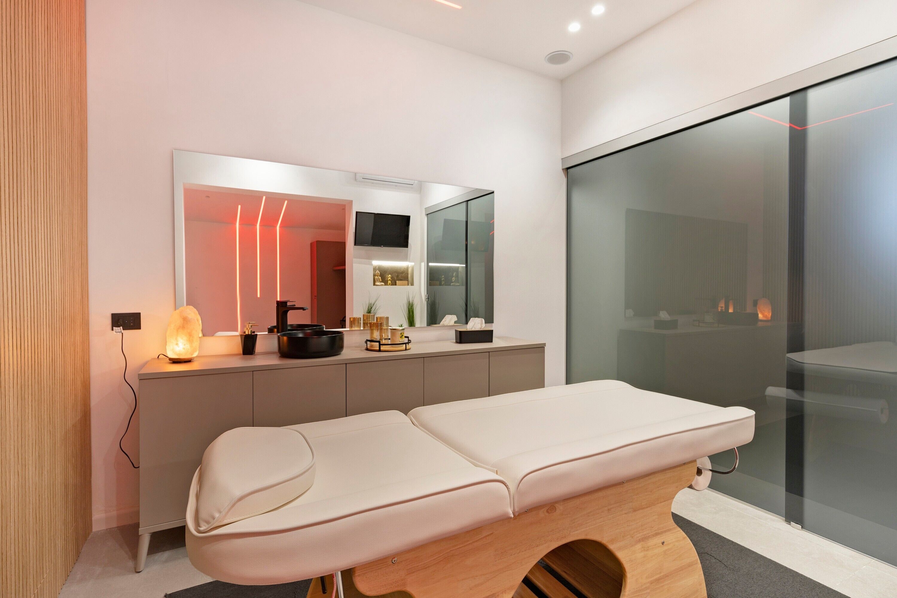 A modern medical examination room with a wooden examination table, a large mirror, and a glass partition.
