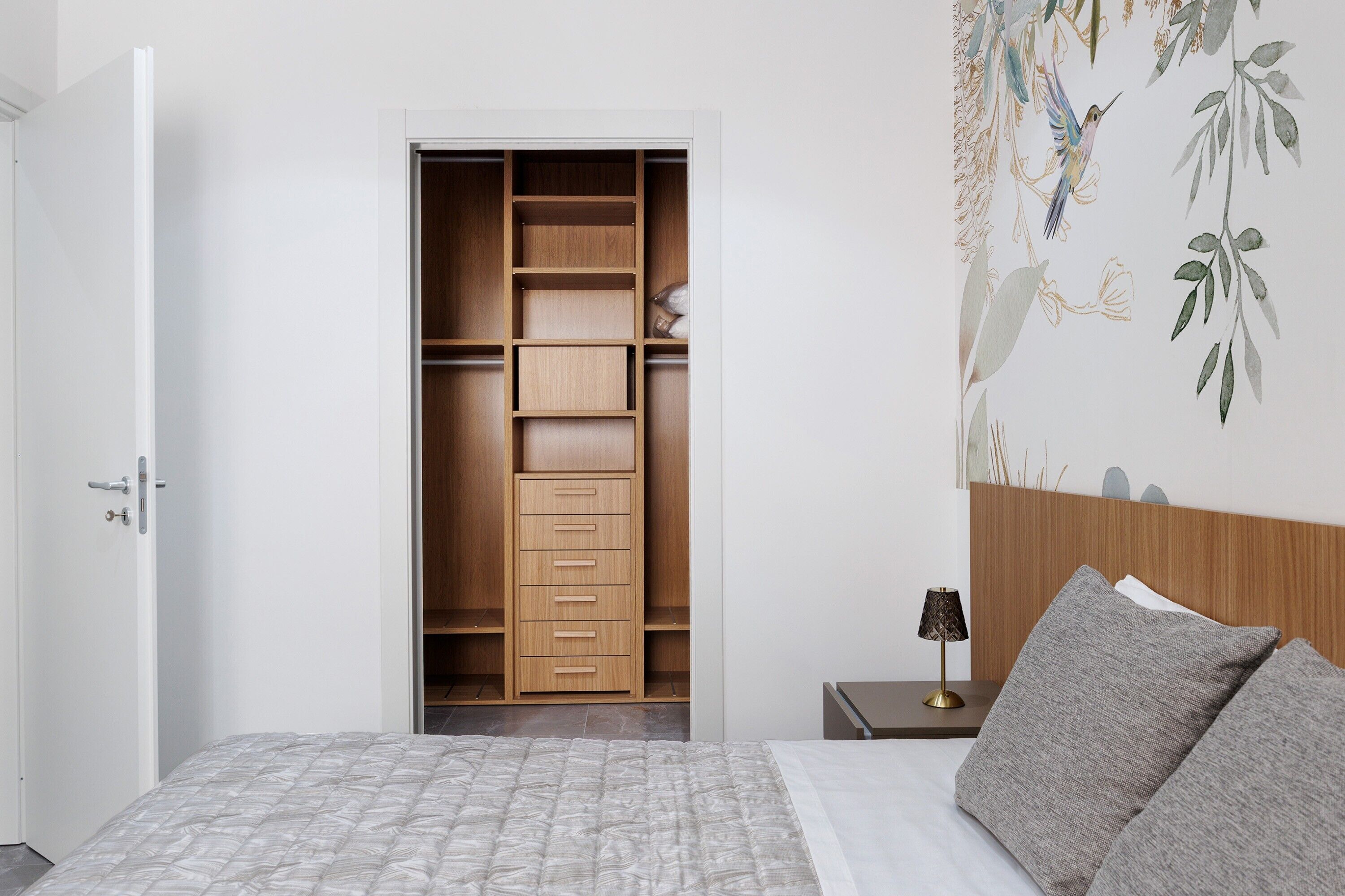 A bedroom with a bed, a nightstand, a wooden wardrobe, and a wall with a nature-themed mural.