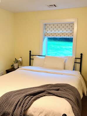 A bedroom with a bed, a window with a view of a green landscape, a bedside table with a lamp, and a potted plant.