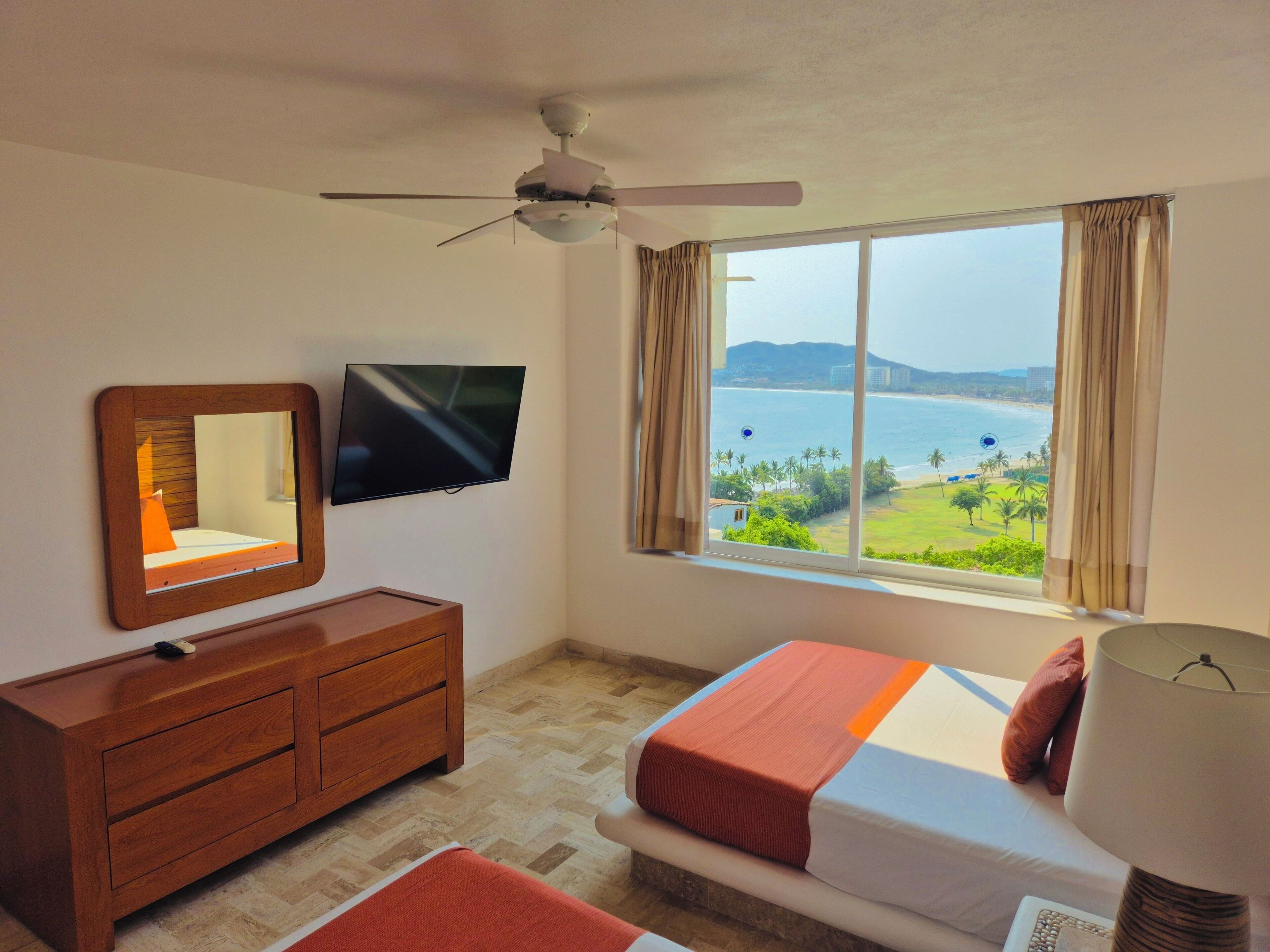 A hotel room with a bed, a TV, a ceiling fan, a dresser, and a window with a view of the sea.
