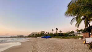 A beach with a sandy shore, palm trees, and a building in the background. - Vista Pacífico • Beachfront Condo • Pool • Ixtapa-Zihuatanejo (Ixtapa Zihuatanejo)